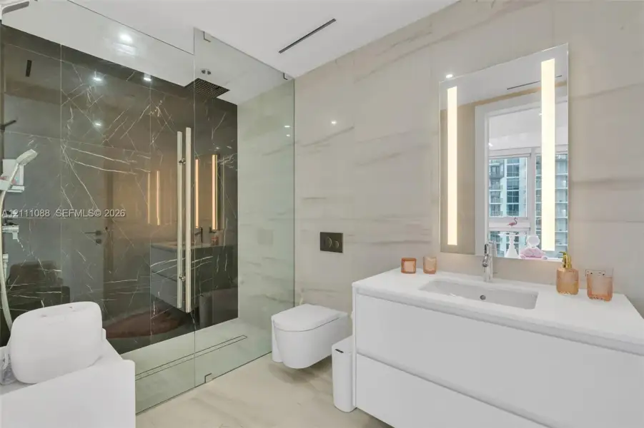 Bathroom featuring large-format light-toned wall tile, glass-enclosed shower with dark contrasting tile, wall-mounted toilet, white double-drawer vanity, and a rectangular mirror with integrated vertical lighting Bathroom featuring large-format light-toned wall tile, glass-enclosed shower with dark contrasting tile, wall-mounted toilet, white double-drawer vanity, and a rectangular mirror with integrated vertical lighting