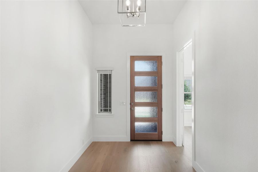 Bright entryway with wood front door, frosted glass panels, hardwood floors, and chandelier overhead.