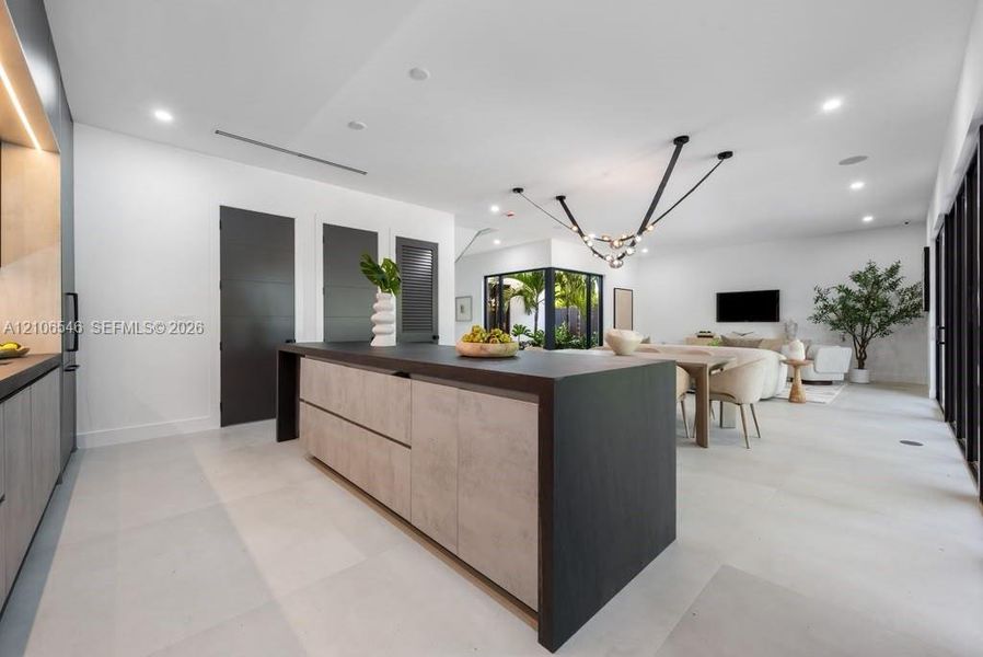 Open-concept living space featuring a central kitchen island with wood-finish cabinetry and a dark countertop, large-format tile flooring, recessed lighting, and a multi-arm modern light fixture
