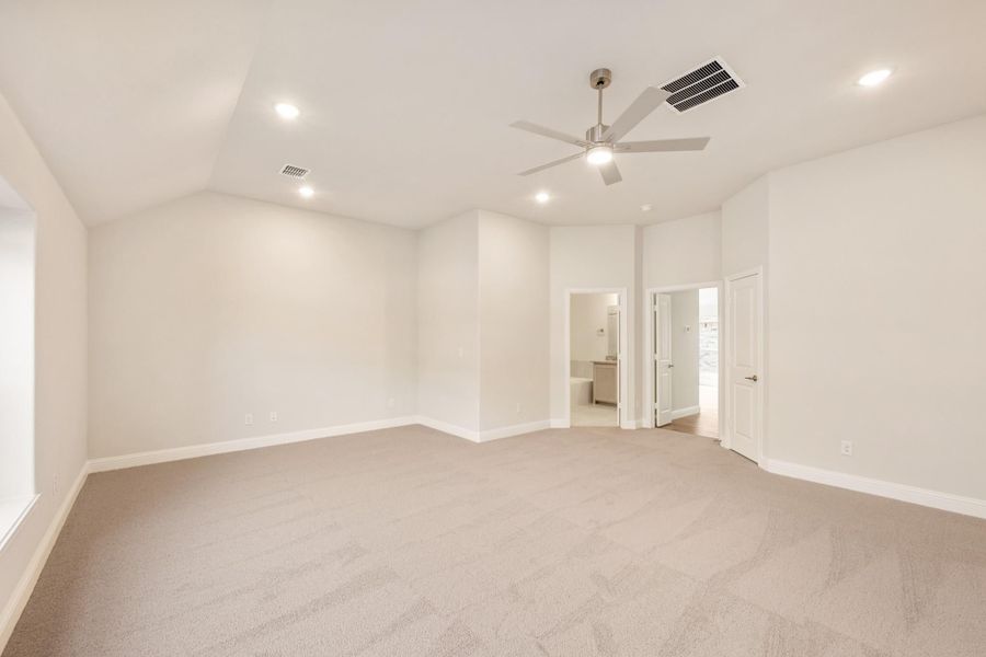 Spacious empty bedroom with vaulted ceiling, ceiling fan, carpet flooring, and doorway to ensuite bathroom Spacious empty bedroom with vaulted ceiling, ceiling fan, carpet flooring, and doorway to ensuite bathroom