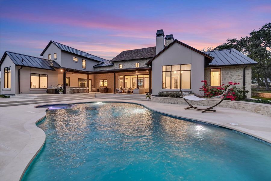 Back of house at dusk featuring a standing seam roof, a metal roof, an outdoor pool, a patio area, and a chimney Back of house at dusk featuring a standing seam roof, a metal roof, an outdoor pool, a patio area, and a chimney