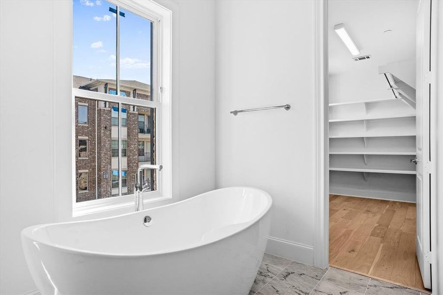 Freestanding soaking tub with chrome fixtures, large window, tiled flooring, white walls, and an adjacent built-in shelving unit with wood-finish flooring