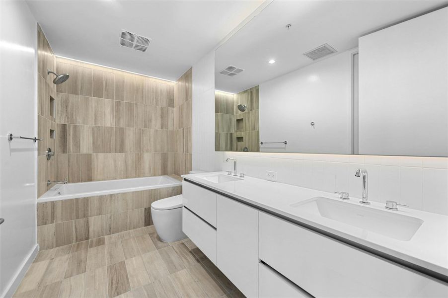 The primary bathroom showcases a dual vanity with sleek white cabinets, providing ample storage & a modern aesthetic plus a large horizontal mirror, enhancing the spacious feel and functionality of the area. The shower and tub combo is designed with floor to ceiling tiles.