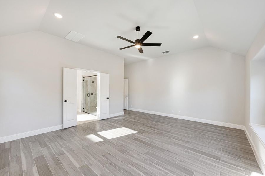 Empty master bedroom with vaulted ceiling, ceiling fan, wood-look tile floors, and open ensuite bathroom door Empty master bedroom with vaulted ceiling, ceiling fan, wood-look tile floors, and open ensuite bathroom door