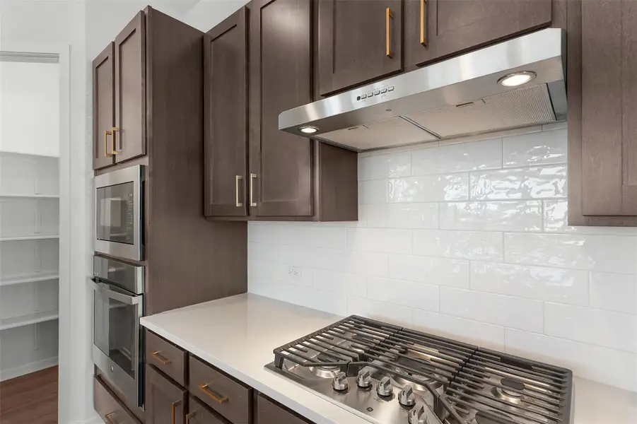 Kitchen with under cabinet range hood, stainless steel appliances, backsplash, dark brown cabinetry, and light stone counters