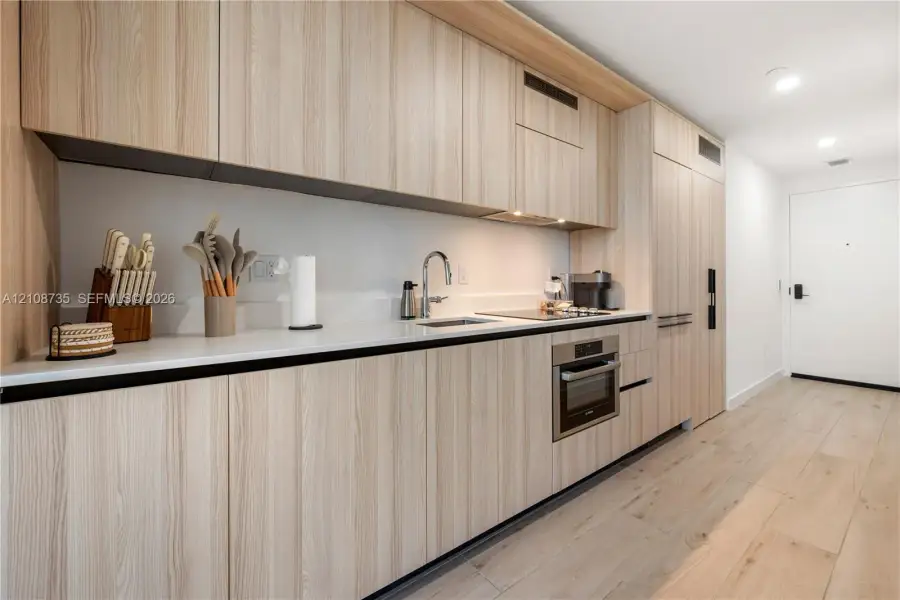 Modern kitchen featuring light wood-finish cabinetry, solid surface countertops, built-in stainless steel appliances, and wide-plank wood-finish flooring Modern kitchen featuring light wood-finish cabinetry, solid surface countertops, built-in stainless steel appliances, and wide-plank wood-finish flooring