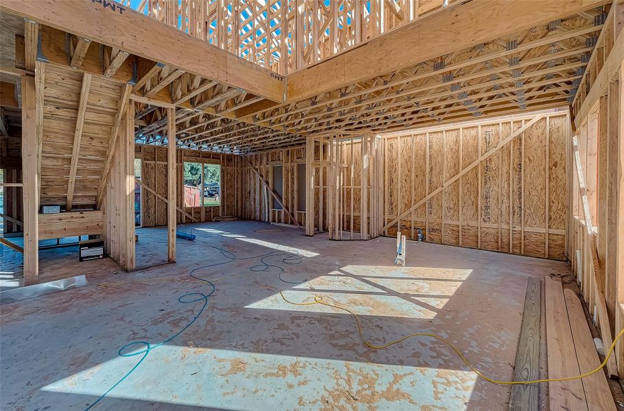This photo shows a partially constructed home interior with exposed wood framing, open spaces, and visible ceiling beams. There are large windows that bring in natural light, and a staircase is visible on the left. The area is wired and ready for further construction.