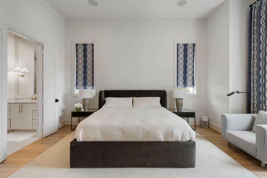 Elegant primary suite designed with balance and symmetry, featuring a refined upholstered bed framed by custom nightstands and designer lighting. Tall ceilings and clean architectural lines create an airy, tranquil atmosphere, while the open entry reveals the beautifully finished en suite bathroom just beyond.