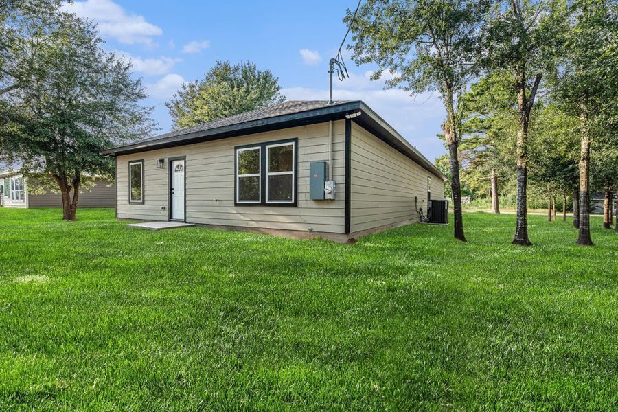 This photo features a modest single-story home with a light-colored exterior and a dark roof. It's surrounded by a spacious, well-maintained green lawn with several trees, providing a serene and private setting. The house has a simple, clean design with two windows and a small entrance walkway.