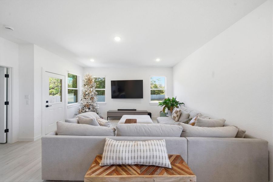 Bright and spacious living room featuring a large sectional sofa, wall-mounted TV, and modern decor. Natural light streams through windows, highlighting a minimalist design with a touch of greenery and holiday decor.