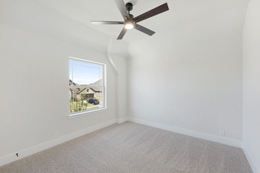 Empty bedroom with vaulted ceiling, ceiling fan, carpet flooring, and a window overlooking the neighborhood.