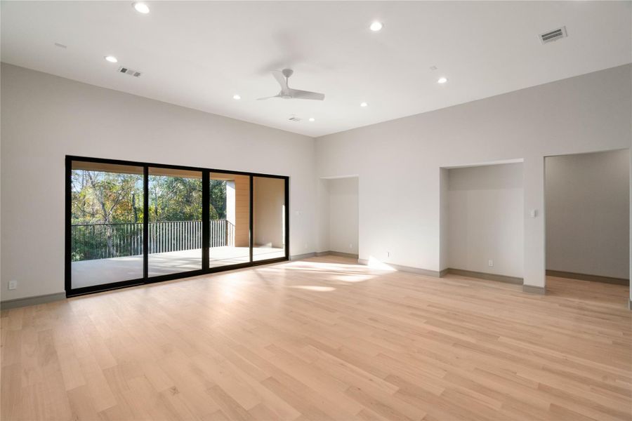 Spacious and bright living area with light wood flooring and recessed lighting. Large glass sliding doors open to a balcony with a scenic view of trees, providing abundant natural light and a seamless indoor-outdoor flow.