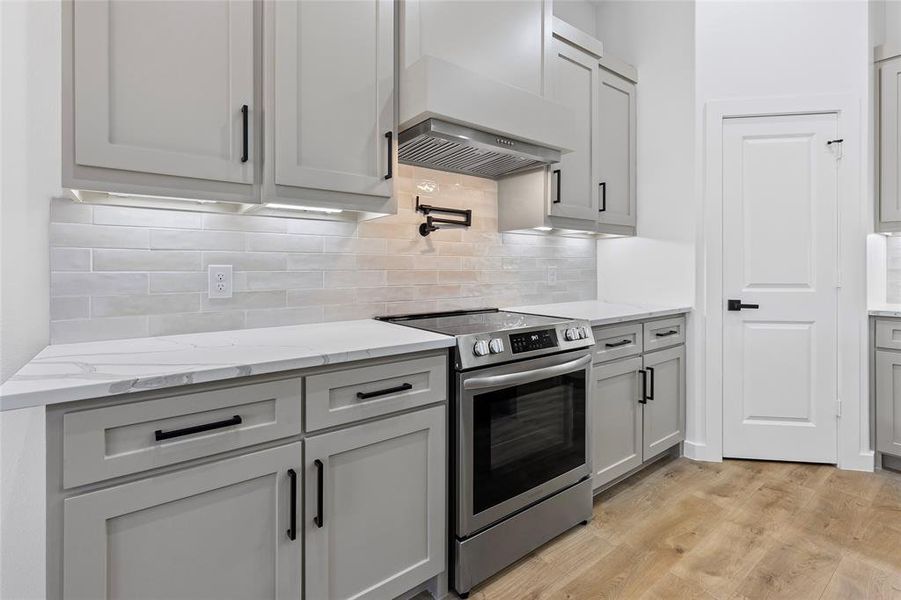 Kitchen featuring light stone counters, custom exhaust hood, stainless steel range with electric cooktop, light wood-style floors, and tasteful backsplash