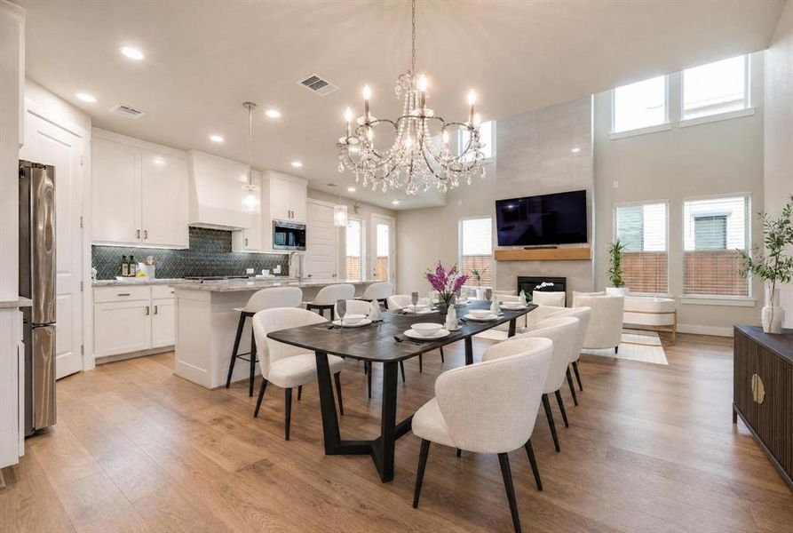 Open-concept living space featuring wood-finish flooring, a contemporary chandelier, a kitchen island with seating, a fireplace with a wood mantle, and high windows providing natural light