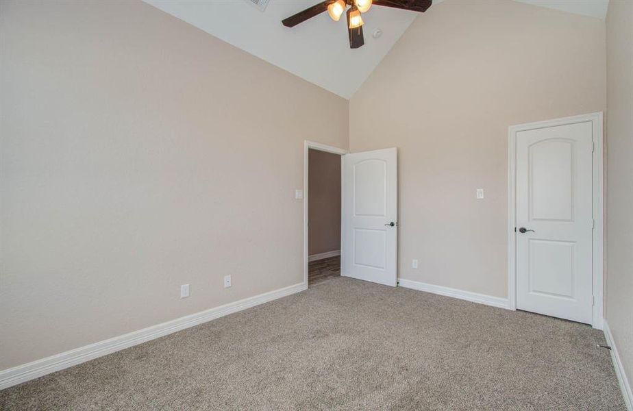 This is a spacious room with high vaulted ceilings, neutral wall paint, and plush carpeting. It features a ceiling fan for comfort and two white doors that likely lead to a closet and an en suite or adjacent room.