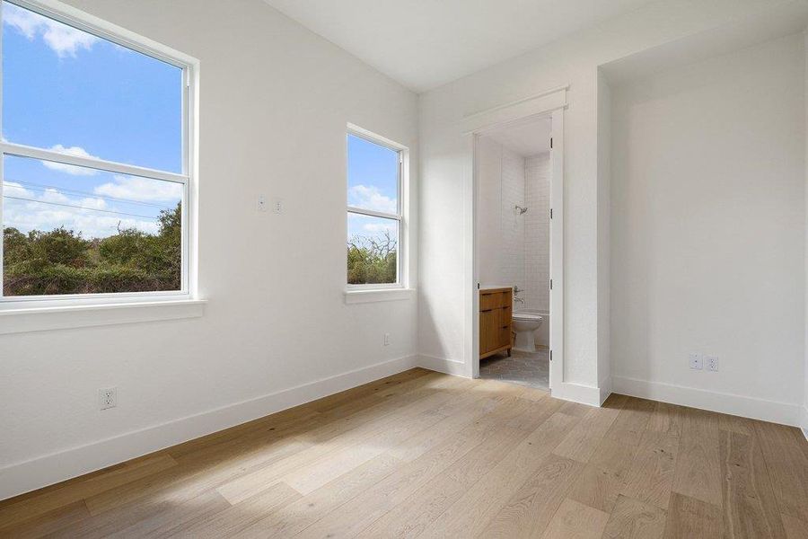 This room features light hardwood flooring, white walls, and two large windows offering views of lush greenery and a clear sky