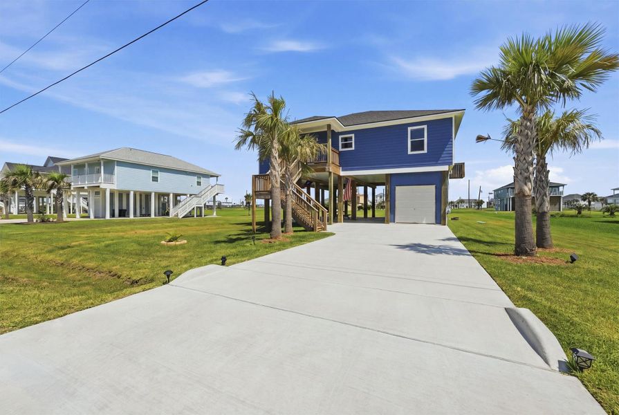 Elevated coastal home with vibrant blue siding, featuring a spacious driveway, lush green lawn, and charming palm trees for a tropical ambiance. Elevated coastal home with vibrant blue siding, featuring a spacious driveway, lush green lawn, and charming palm trees for a tropical ambiance.