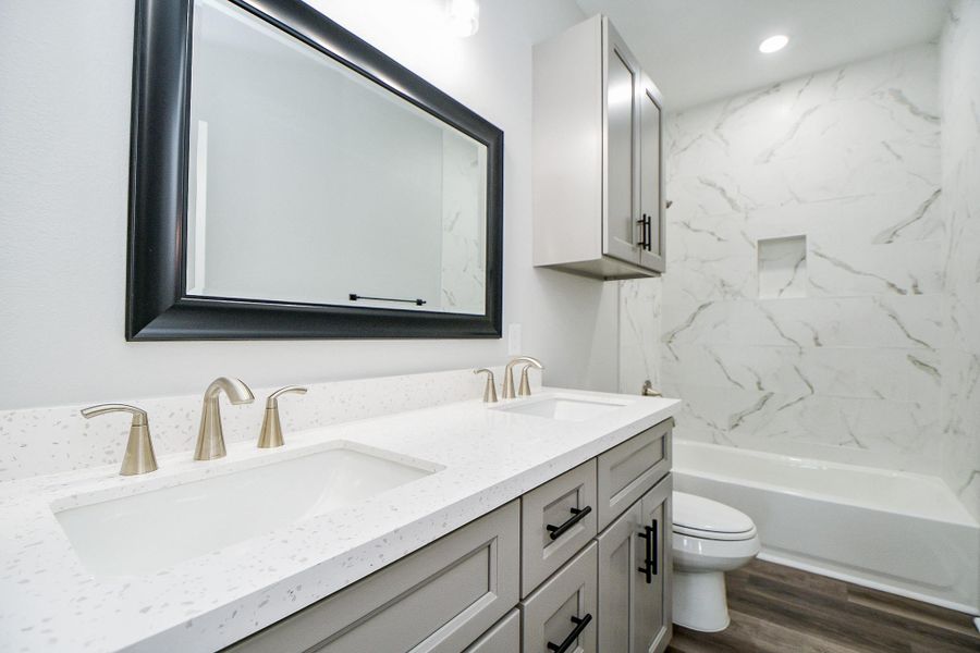 This is a modern secondary bathroom featuring a double vanity with brushed nickel fixtures, white countertops, and a large mirror. The space includes a tub-shower combo with marble-like wall tiles, a toilet, and grey cabinetry offering ample storage. The flooring has a wood-look finish. This is a modern secondary bathroom featuring a double vanity with brushed nickel fixtures, white countertops, and a large mirror. The space includes a tub-shower combo with marble-like wall tiles, a toilet, and grey cabinetry offering ample storage. The flooring has a wood-look finish.