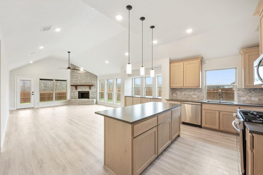 Open-concept kitchen with center island, light wood cabinets, and vaulted ceiling open to living room with stone fireplace