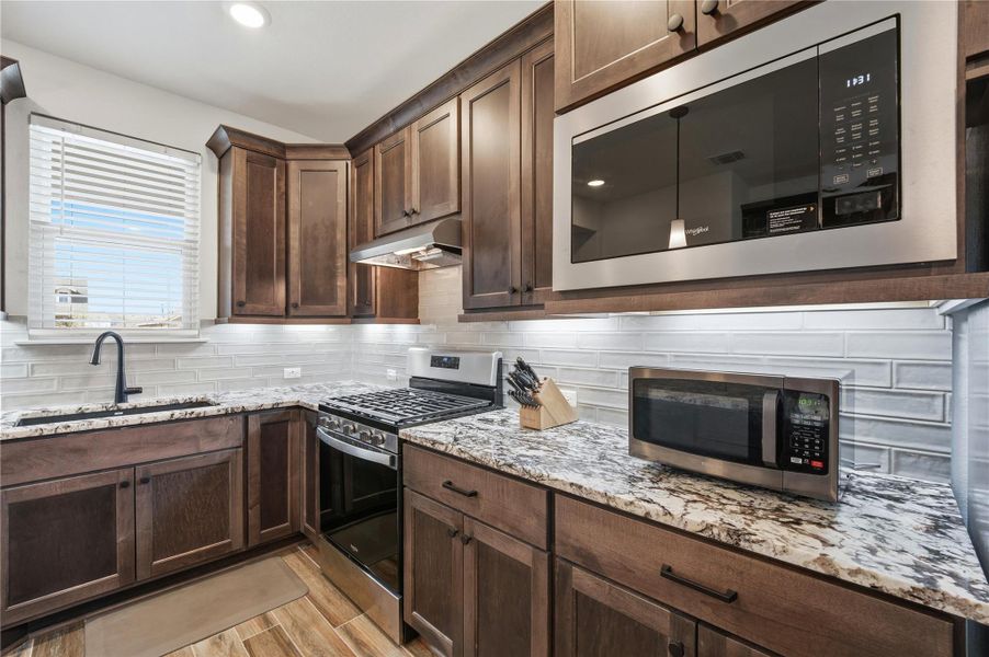 Kitchen featuring stainless steel appliances, dark wood finish cabinets, light stone countertops, tasteful backsplash, and light wood-style floors