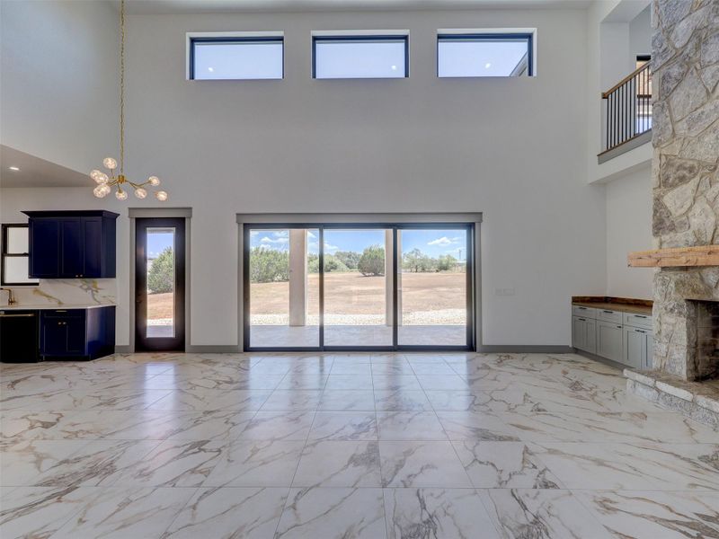 Double-height ceilings and a stone fireplace frame this light-filled room with marble-patterned tile flooring.