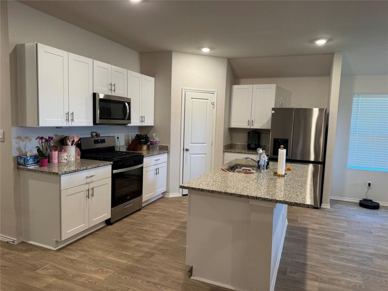 Modern kitchen featuring white cabinetry, granite-style countertops, stainless steel appliances, a center island with sink, and wood-finish flooring