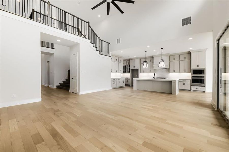Open-concept living space featuring light wood-finish flooring, a modern kitchen with light gray cabinetry and an island, stainless steel built-in appliances, and a staircase with an open black metal railing leading to an upper landing Open-concept living space featuring light wood-finish flooring, a modern kitchen with light gray cabinetry and an island, stainless steel built-in appliances, and a staircase with an open black metal railing leading to an upper landing