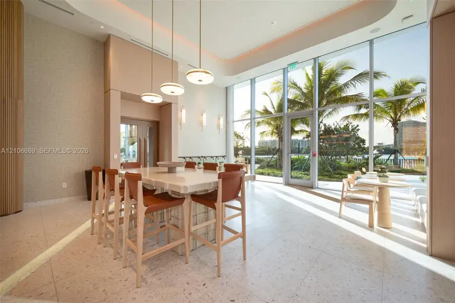 Expansive common area featuring floor-to-ceiling glass windows, recessed tray ceiling lighting, stone-top island with integrated seating, and polished stone flooring