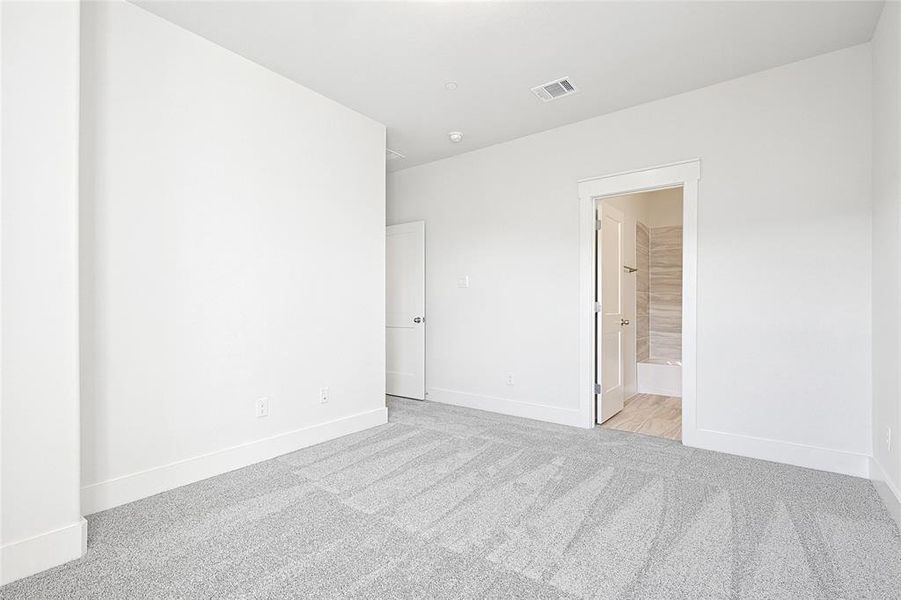 Bedroom featuring light grey carpeting, white walls, and a white interior door, with an open doorway leading to a bathroom with tiled walls and flooring
