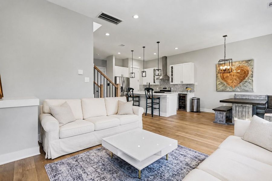 This open-concept living area features a modern kitchen with white cabinetry and a stylish backsplash, seamlessly connected to a cozy living space with green leather sofas. Hardwood floors and contemporary lighting add warmth and elegance.
