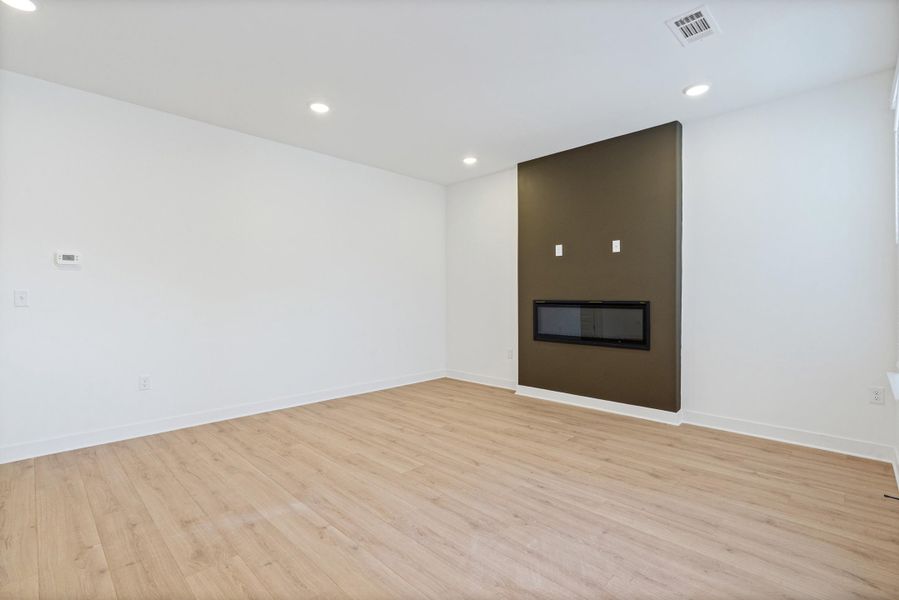 Modern living space featuring a linear electric fireplace, contrasting accent wall, wood-finish flooring, recessed lighting, and white baseboards Modern living space featuring a linear electric fireplace, contrasting accent wall, wood-finish flooring, recessed lighting, and white baseboards