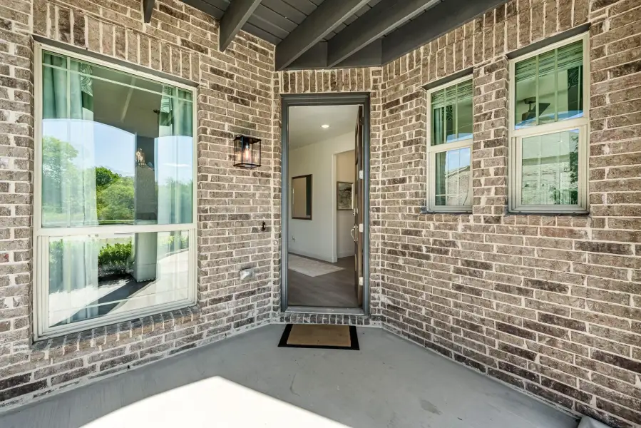 Home entry with brick exterior, open front door, wall lantern, and welcome mat on covered porch