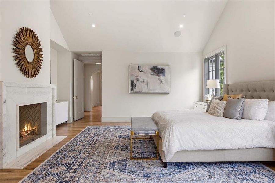 Vaulted ceiling bedroom featuring a fireplace with a stone surround, hardwood-style flooring, a large window, and an arched hallway entry Vaulted ceiling bedroom featuring a fireplace with a stone surround, hardwood-style flooring, a large window, and an arched hallway entry