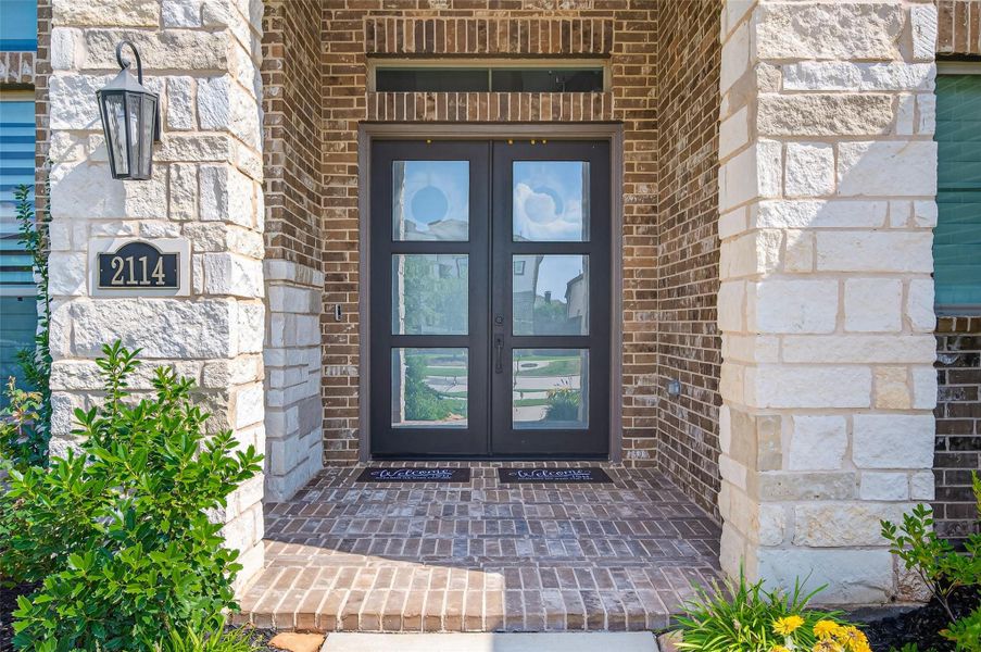 This photo shows a stylish and welcoming front entrance with double glass doors framed by brick and stonework. The porch is accented with greenery and a modern light fixture, creating an inviting first impression. This photo shows a stylish and welcoming front entrance with double glass doors framed by brick and stonework. The porch is accented with greenery and a modern light fixture, creating an inviting first impression.