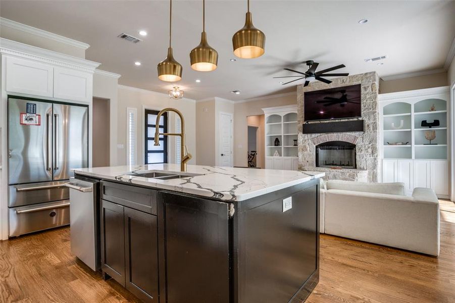 Kitchen with light stone countertops, stainless steel appliances, a stone fireplace, hanging light fixtures, and an island with sink