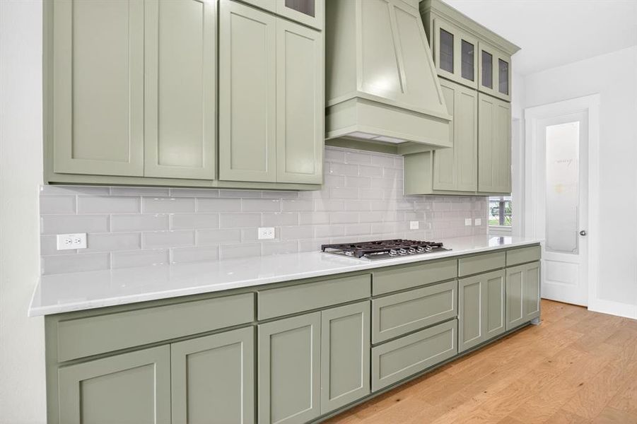 Kitchen featuring green shaker cabinetry, white countertops, a gas range with a matching range hood, a subway tile backsplash, and wood-finish flooring Kitchen featuring green shaker cabinetry, white countertops, a gas range with a matching range hood, a subway tile backsplash, and wood-finish flooring