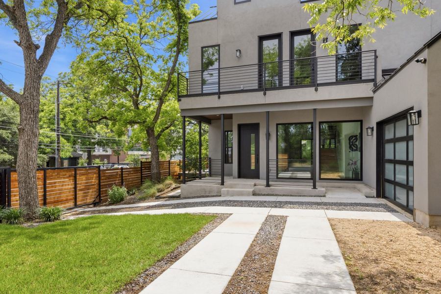 Modern stucco exterior featuring a second-story balcony with metal railing, an integrated garage with a full-view door, a concrete and gravel driveway, a wood fence, and mature trees