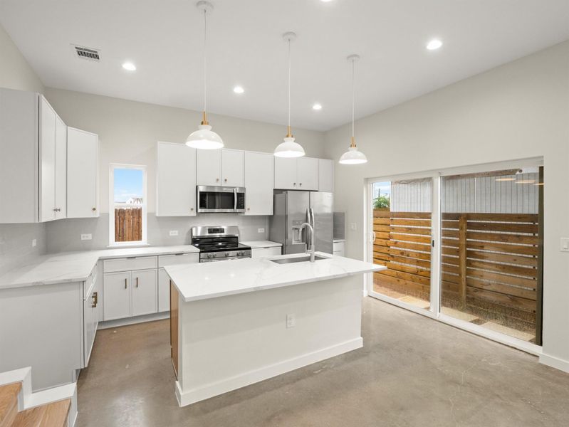 Modern kitchen featuring white cabinetry, stainless steel appliances, a center island with sink, concrete flooring, and a sliding glass door to a privacy-fenced outdoor area
