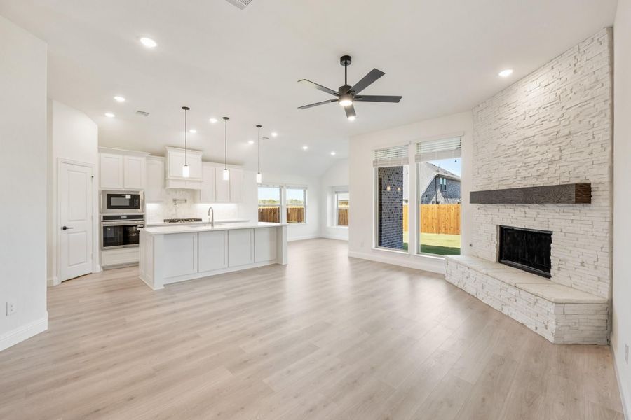 Open-concept living area with stone fireplace, light wood floors, and white kitchen with island and pendant lights Open-concept living area with stone fireplace, light wood floors, and white kitchen with island and pendant lights