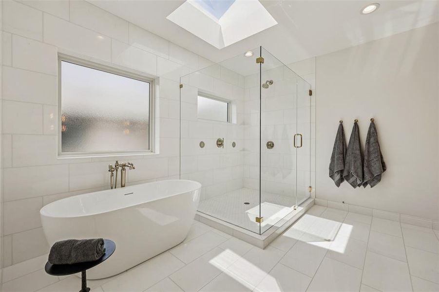 Freestanding soaking tub with chrome fixtures, glass-enclosed shower with brass accents, white tiled walls, and a recessed ceiling light