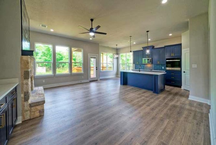 A vibrant kitchen with blue cabinetry and an island flows seamlessly into a well-lit living area featuring large windows and a stone fireplace.