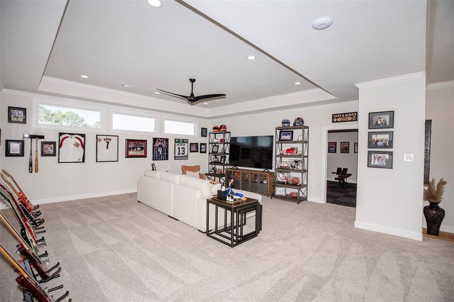 This spacious entertainment room features carpeted flooring, a large TV, and wall displays for sports memorabilia. It includes a cozy seating area and ample natural light from high windows, perfect for relaxing or hosting gatherings.