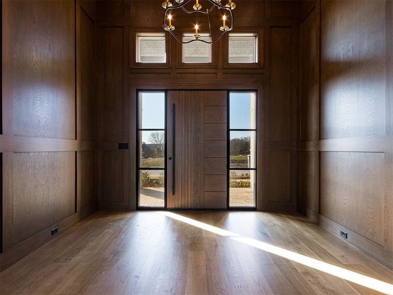 Light and bright foyer entrance with wood door and flooring, a designer chandelier, and surrounded in white oak paneling.