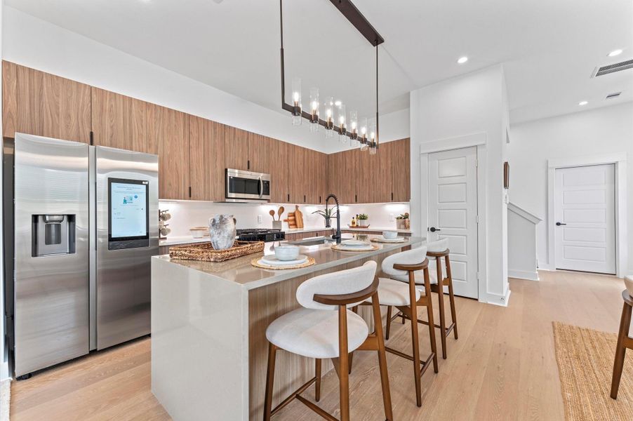 Striking and modern kitchen island serves as the centerpiece of this open-concept culinary space, featuring a waterfall quartz countertop that flows seamlessly down one side for a sculptural, high-end look. The island is framed by minimalist cabinetry in matte finishes, paired with bold pendant lighting that adds a touch of sophistication. Integrated bar seating invites casual dining and conversation, while the clean lines and neutral palette enhance the island’s sleek aesthetic.