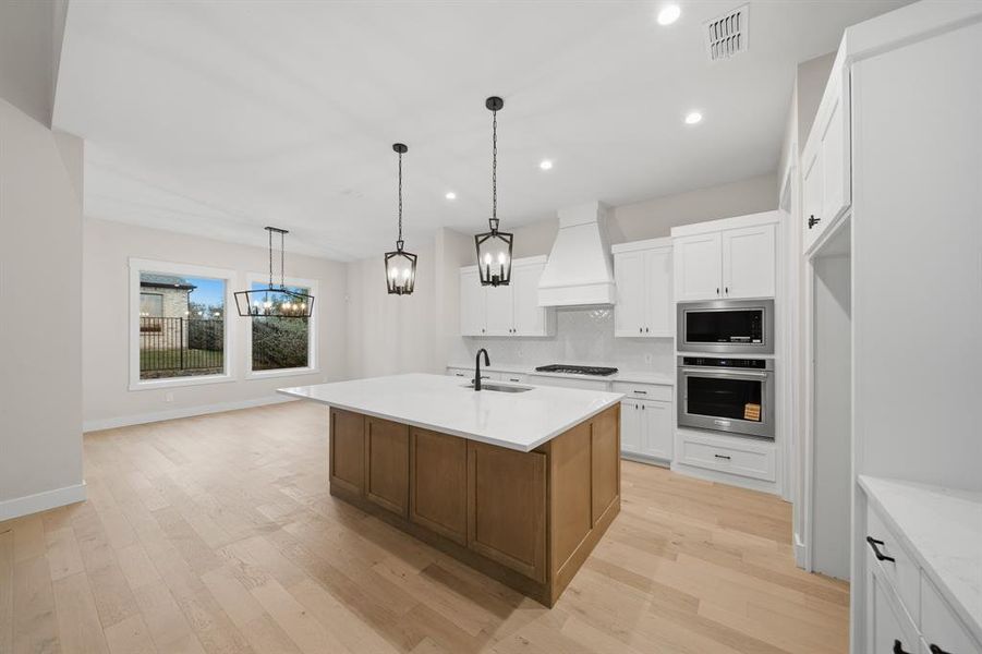 Kitchen with white cabinets, hanging light fixtures, an island with sink, light wood finished floors, and stainless steel appliances