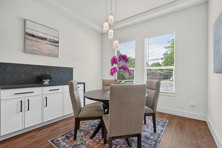 Bright dining area featuring wood-finish flooring, a contemporary chandelier, two windows with blinds, and white built-in cabinetry with a dark countertop and backsplash