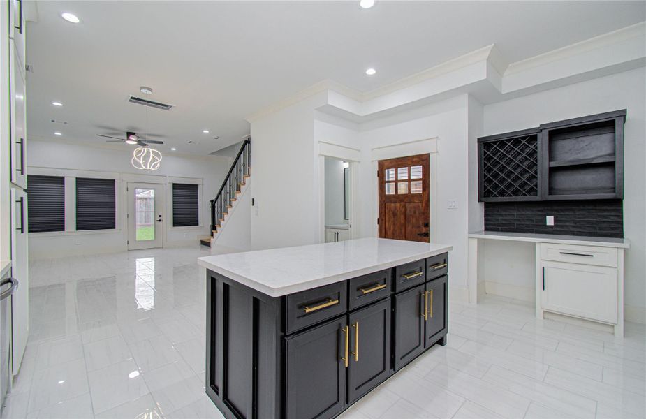 Home features a bright open-concept layout with sleek white walls and glossy tile flooring. The spacious kitchen includes a central island with dark cabinetry and gold accents, complementing the elegant wine rack and sleek design. The living area has large windows, a stylish light fixture, and access to an outdoor covered patio. A staircase leads to the upper level, enhancing the home's contemporary feel.