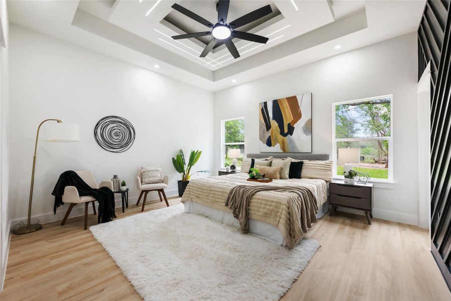 Spacious bedroom featuring light wood-finish flooring and a multi-tiered tray ceiling with recessed lighting and a ceiling fan