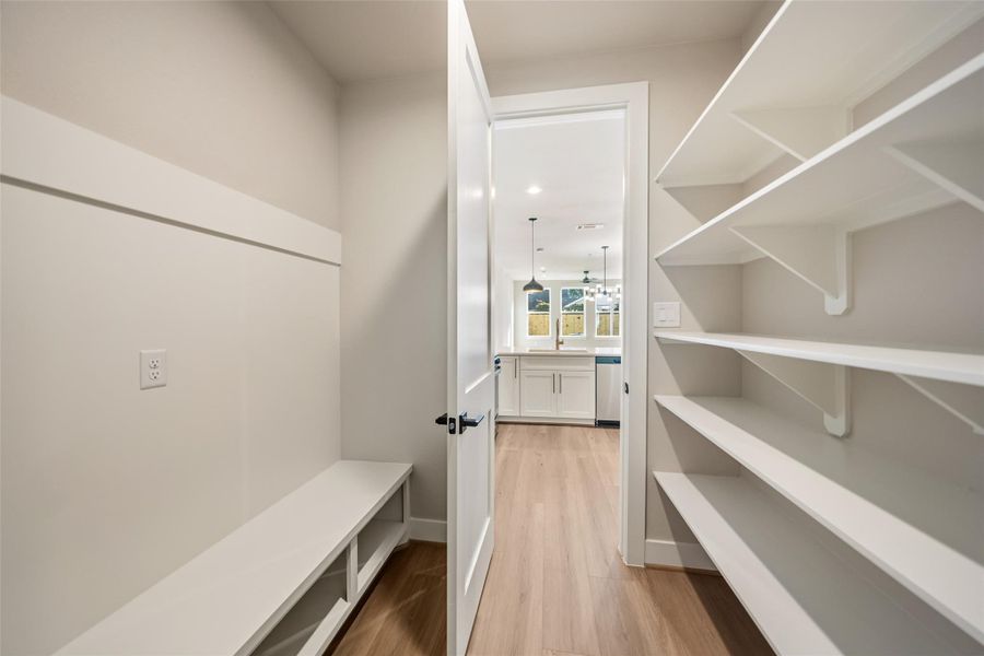 This photo showcases a bright pantry or storage area with white shelving on one side and a bench with storage underneath on the other. The space leads into a kitchen with modern pendant lighting and a view of large windows, providing a spacious and open feel.