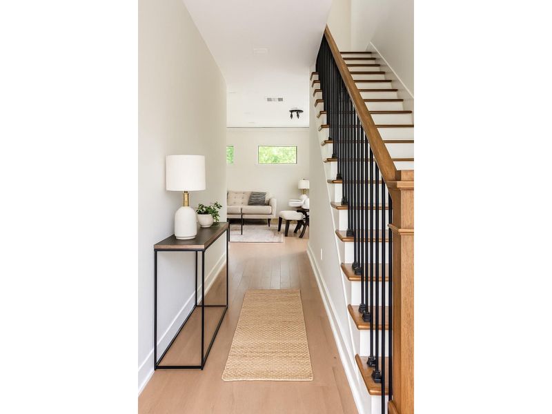 Bright and inviting entry hallway with clean modern finishes, light wood flooring, and an elegant staircase with wood treads and black iron balusters, leading to an open living space filled with natural light.